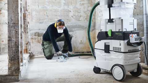 Handwerker kniet auf dem Boden und schleift ihn mit einem Festool Renofix Schleifer. Daneben steht ein Festool Absaugmobil.